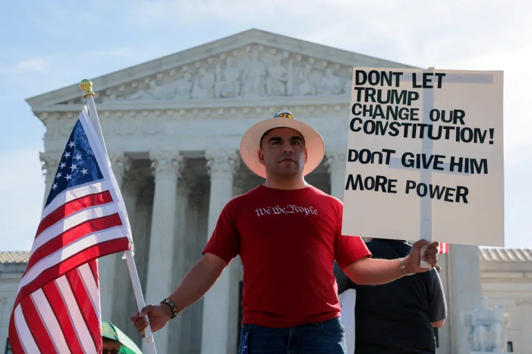 En demonstrant iført en T-skjorte med uttrykket "Vi folket" holder et skilt og et amerikansk flagg utenfor den amerikanske høyesterettsbygningen den dagen retten forventes å høre muntlige argumenter om lovligheten av Trump-administrasjonens innsats for å begrense fødselsrettsborgerskap for barn av immigranter, i Washington, DC, USA, 1. april 2026. REUTERS/Kylie Cooper TPX DAGENS BILDER