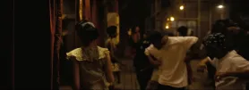 En svakt opplyst innendørsscene med flere mennesker samlet, noen danser og andre observerer.