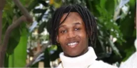 En smilende person med dreadlocks iført en hvit turtleneck-genser mot en uskarp grønn bakgrunn.